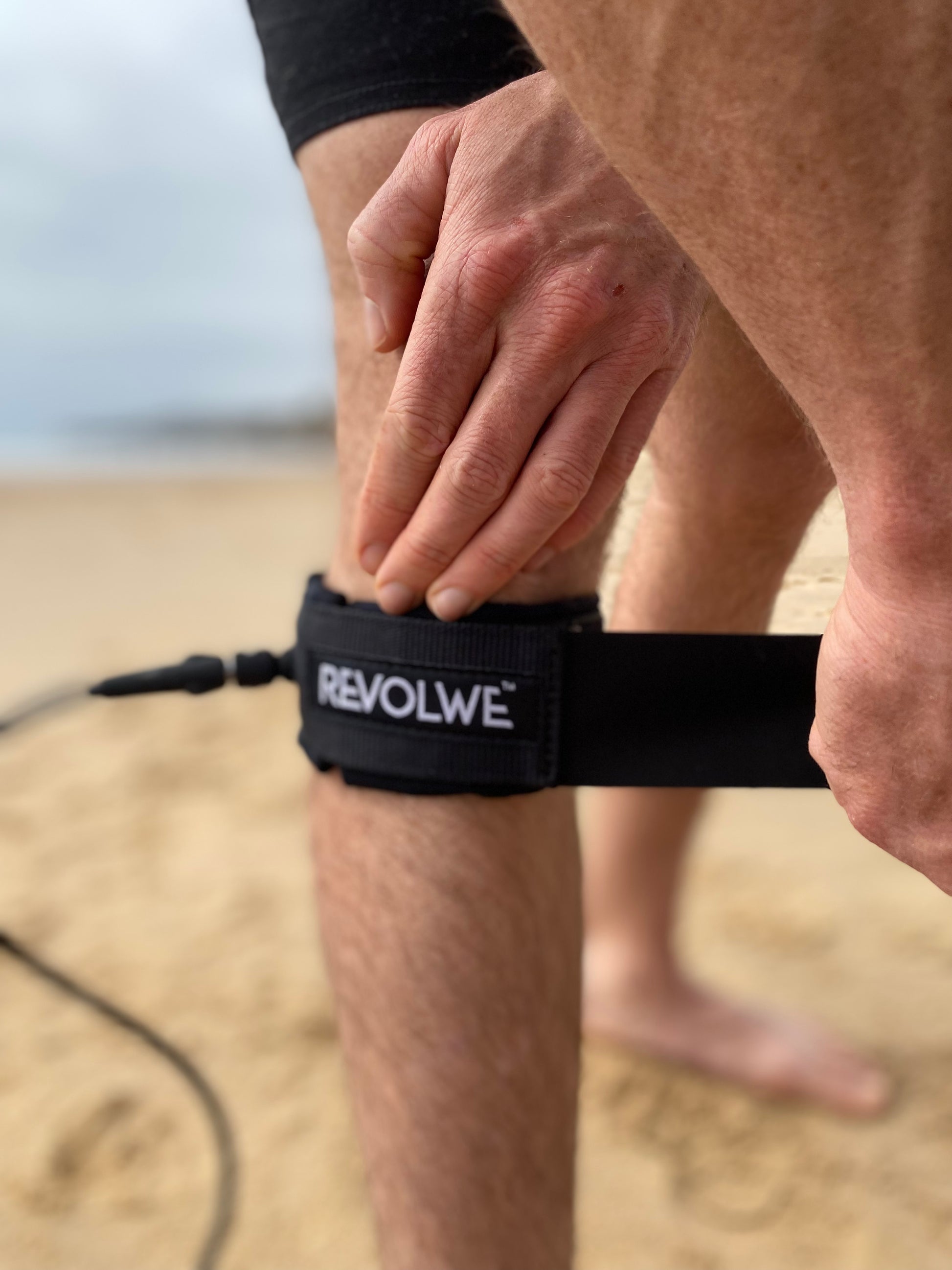 A closeup of a surf leash cuff being put onto a leg.The brand name REVOLWE is on the cuff. The beach and sea are visible in the background.